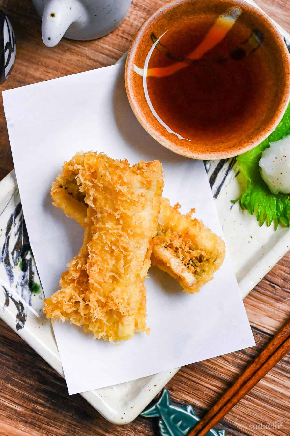 two pieces of conger eel tempura on a rectangular plate with tempura dipping sauce and grated daikon on a perilla leaf