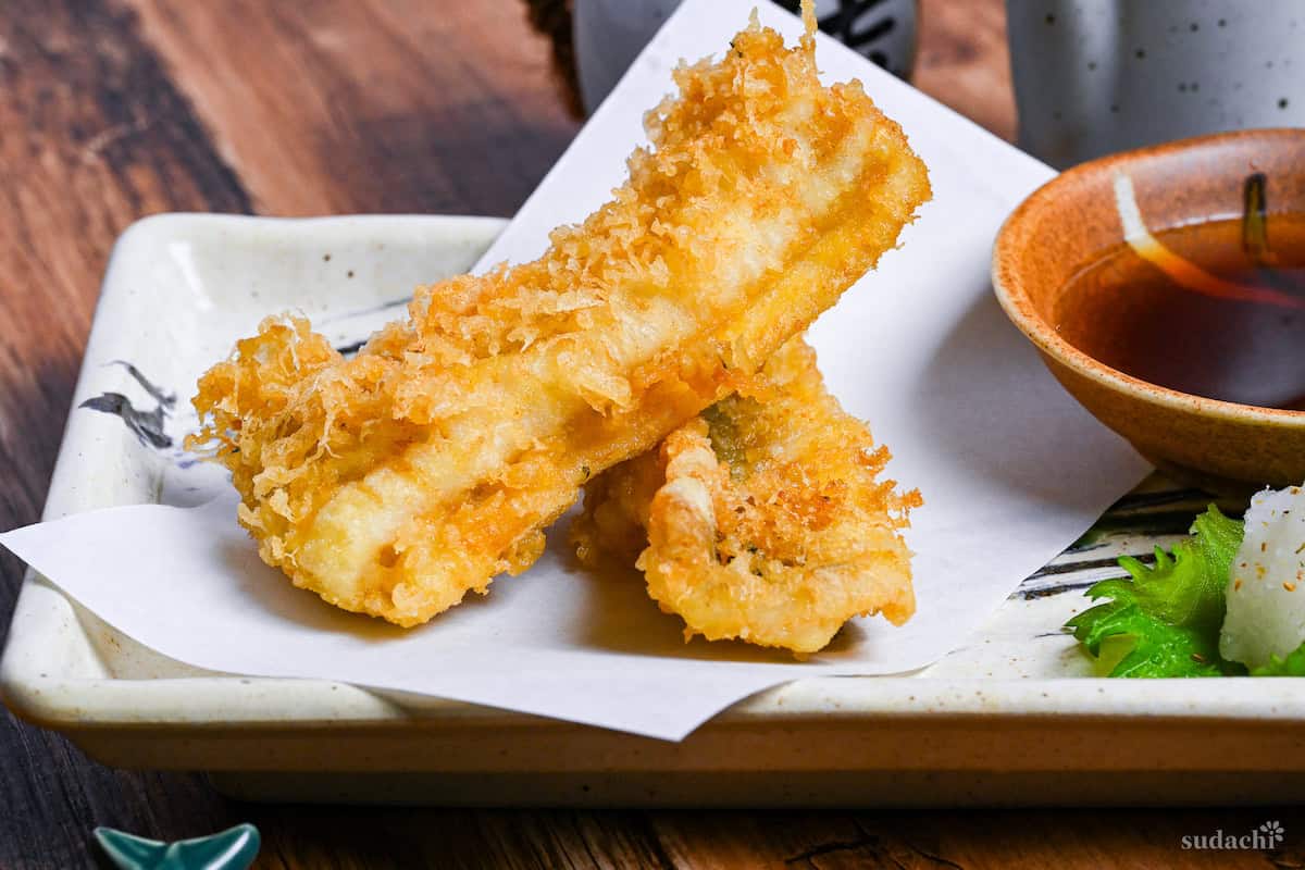 two pieces of conger eel tempura on a rectangular plate with tempura dipping sauce and grated daikon on a perilla leaf