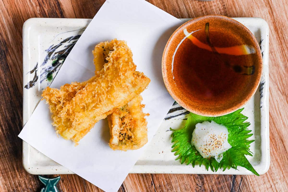 two pieces of conger eel tempura on a rectangular plate with tempura dipping sauce and grated daikon on a perilla leaf top down