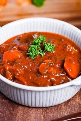 Japanese style beef stew made with tomatoes and mushrooms in a white ceramic pot with small handles next to a glass of red wine and sliced toasted baguette on wooden table