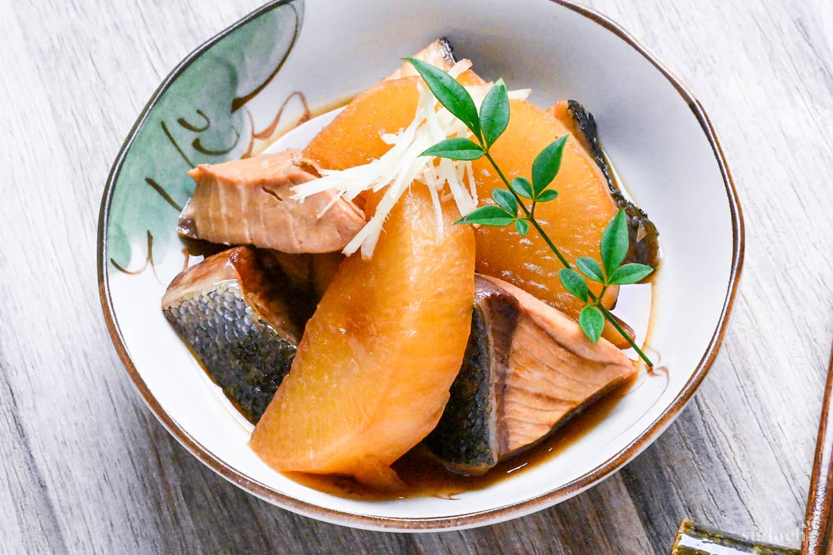 Buri Daikon (Simmered Yellowtail and Daikon Radish) in a small cream bowl with Japanese design topped with decorative leaves