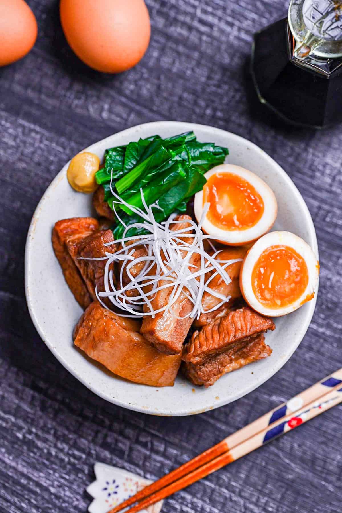 Buta no kakuni (Japanese braised pork belly) in an off-white dish with marinated soft boiled eggs, mustard spinach and a blob of mustard topped with shredded green onion