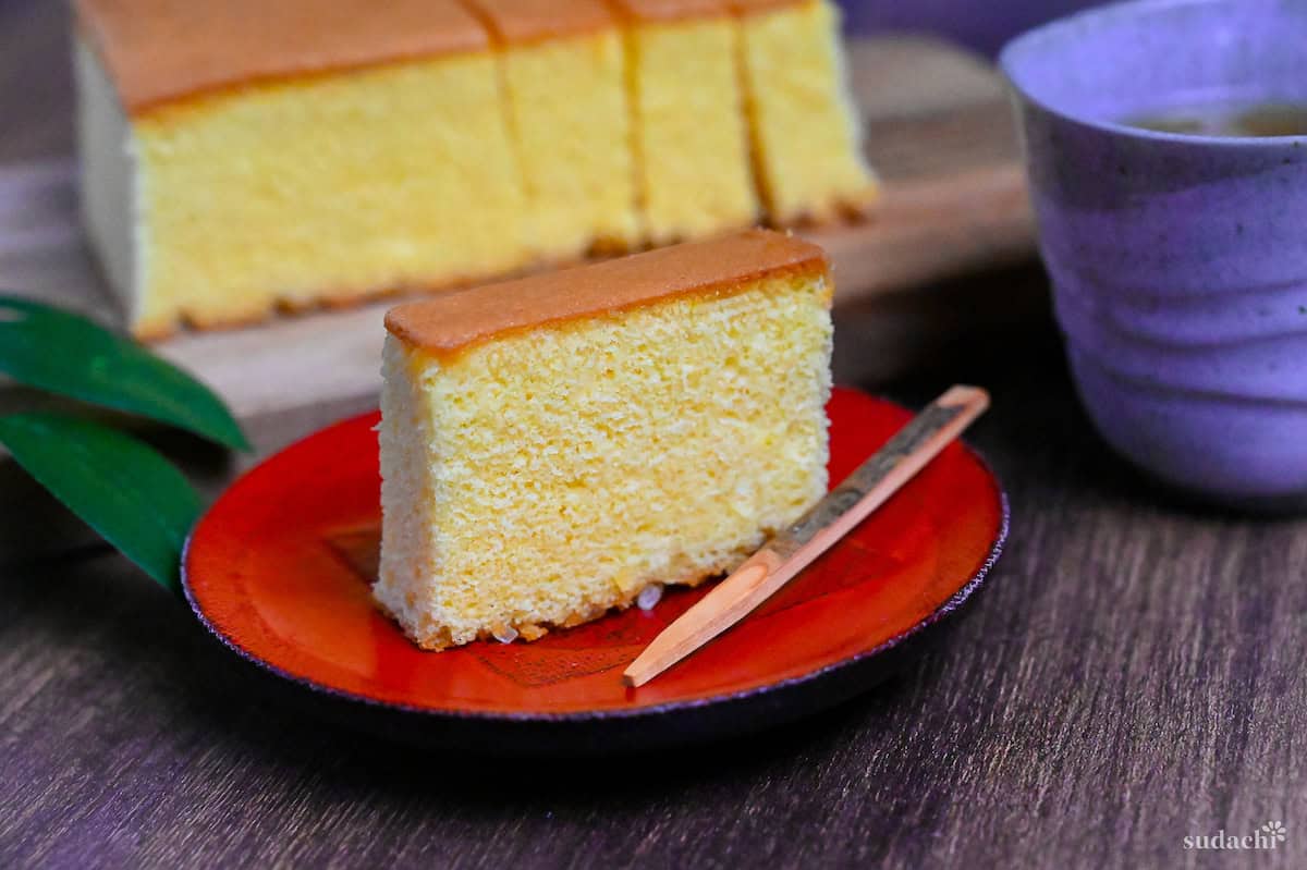 A slice of castella cake on a red plate with a more slices cut on a wooden chopping board in the background