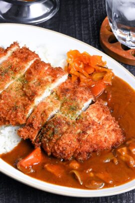 Japanese chicken katsu curry served on a white oval-shaped plate with beige rim, next to a wooden spoon on a black background