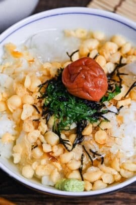 Chilled ume chazuke topped with tempura flakes, bonito flakes, kizami nori, shredded shiso leaves and one umeboshi in a white and blue flower-design bowl