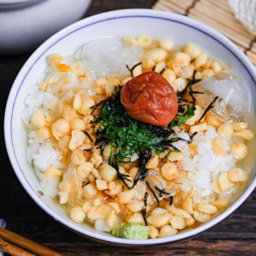Chilled ume chazuke topped with tempura flakes, bonito flakes, kizami nori, shredded shiso leaves and one umeboshi in a white and blue flower-design bowl