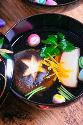 Japanese Clear Soup (Osuimono) in a black bowl with decorative lid, made with shiitake mushroom, kamaboko fish cake, dried wheat gluten (temari fu), yuzu peel and Japanese wild parsley