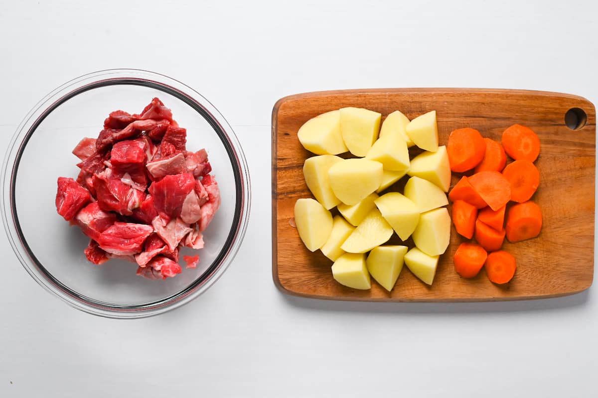 Beef cut into bitesize cubes in a glass bowl and potato and carrots roughly cut on a wooden chopping board