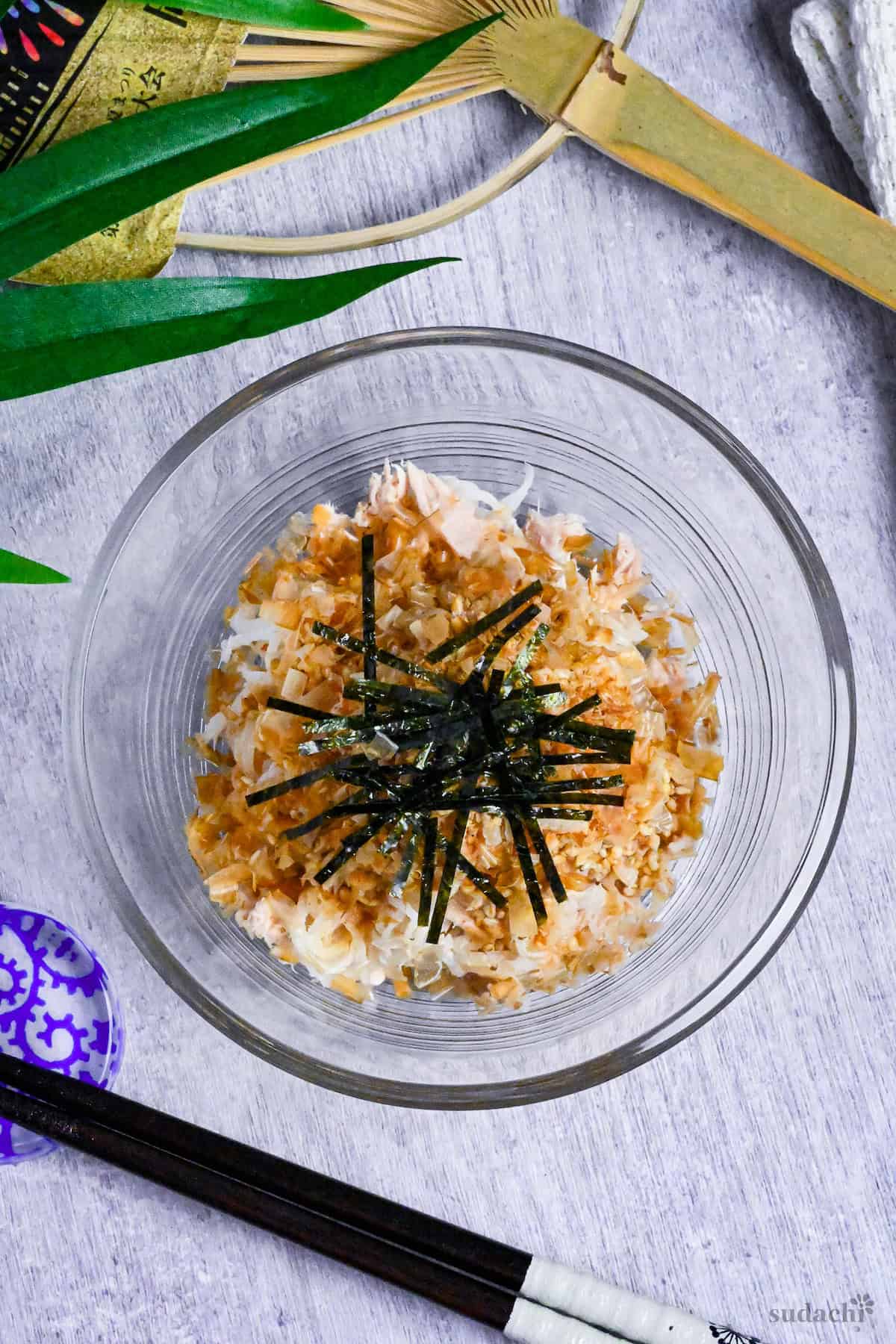 Addictive daikon radish salad topped with bonito flakes, sesame seeds and kizami nori served in a glass bowl next to black chopsticks and a Japanese fan in the background