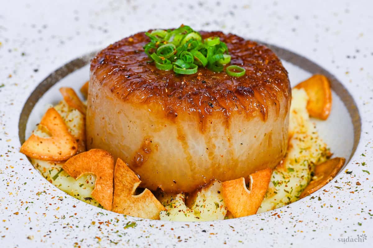 A daikon steak served over mashed potato and sprinkled with crispy garlic chips on a white plate with gold rim side view