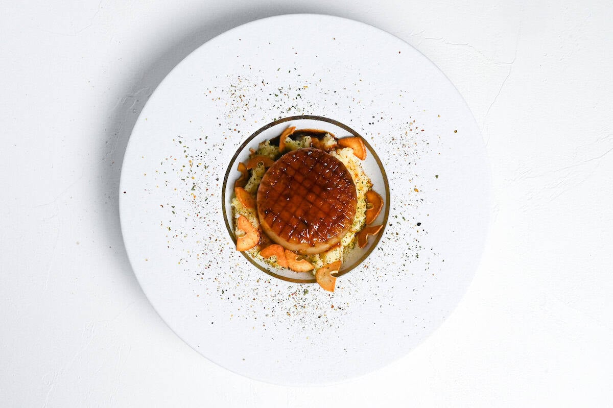 A daikon steak served over mashed potato and sprinkled with crispy garlic chips on a white plate with gold rim