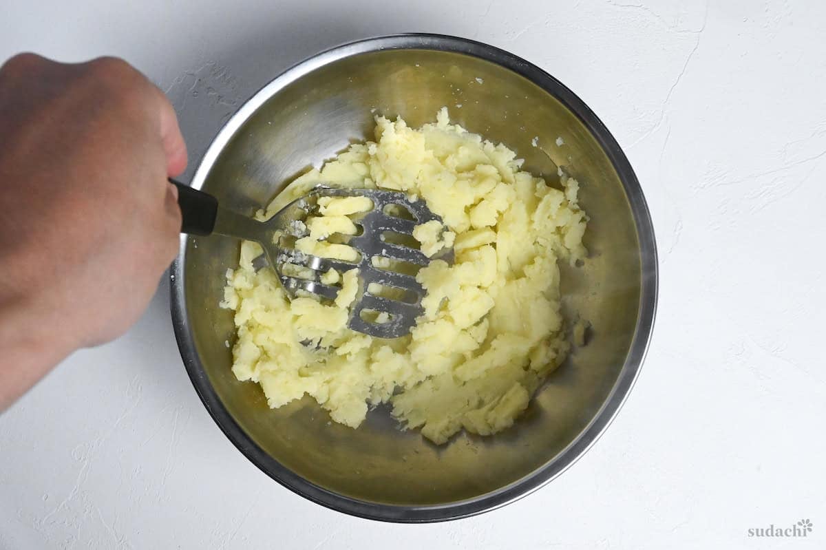 mashing potatoes in a steel mixing bowl with a metal potato masher