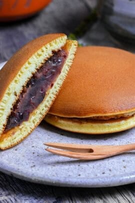 Half a dorayaki pancake revealing tsubuan (chunky red bean paste) in the center resting on a whole dorayaki on a mottled plate