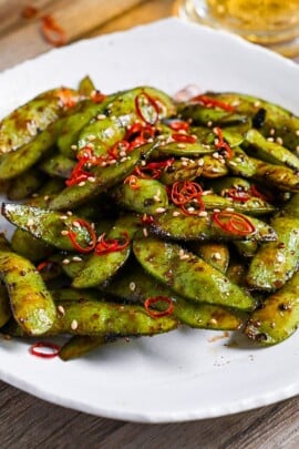 Stir fried edamame with garlic and oyster sauce glaze topped with dried chili and sesame seeds on a white plate