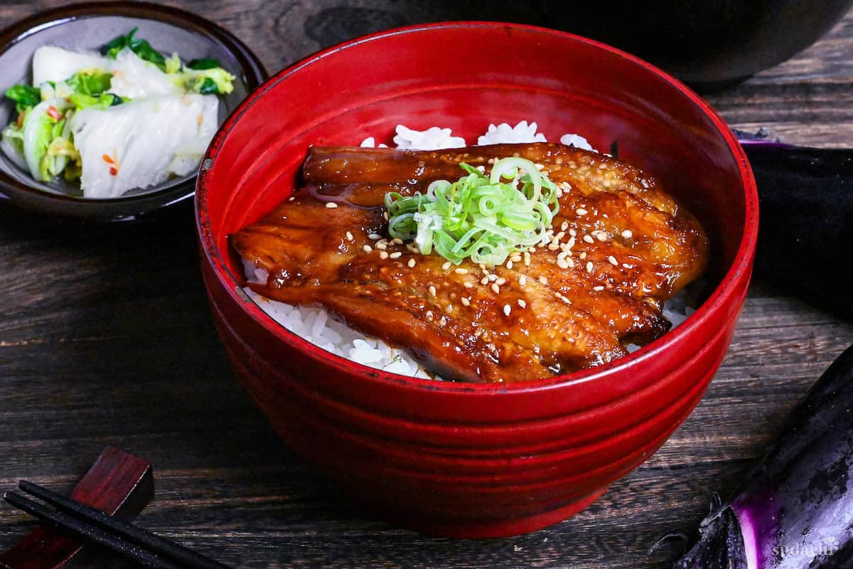 eel kabayaki in a red bowl on a dark wooden surface surrounded by raw eggplants, napa cabbage picjles and clear soup