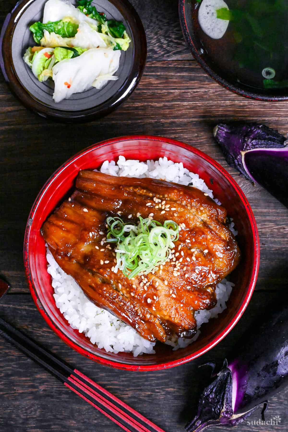 eel kabayaki in a red bowl on a dark wooden surface surrounded by raw eggplants, napa cabbage picjles and clear soup