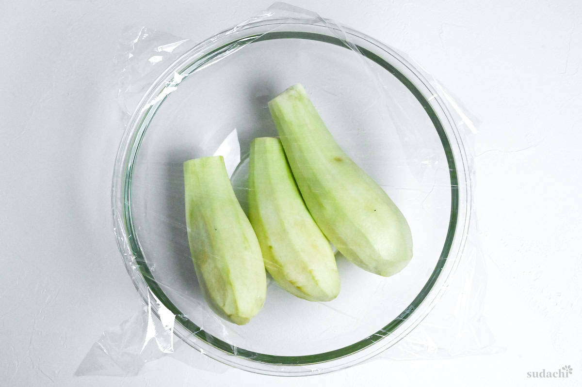 peeled eggplants in a glass bowl covered with plastic wrap ready for microwaving