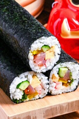 3 ehomaki (lucky direction sushi rolls) on a bamboo platform with a box of soy beans, an oni mask and sushi oke filled with sushi rice in the background