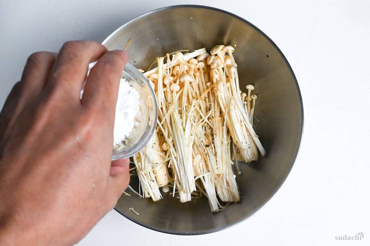 a hand holding a small glass bowl of potato starch above seasoned enoki mushrooms in a mixing bowl
