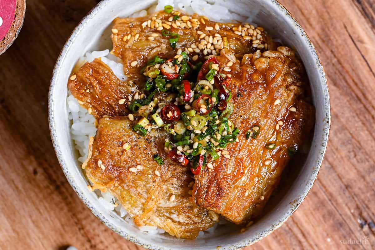 Crispy enoki mushroom rice bowl topped with green onions, sesame seeds and dried red chilis in an off white bowl on a wooden surface top down
