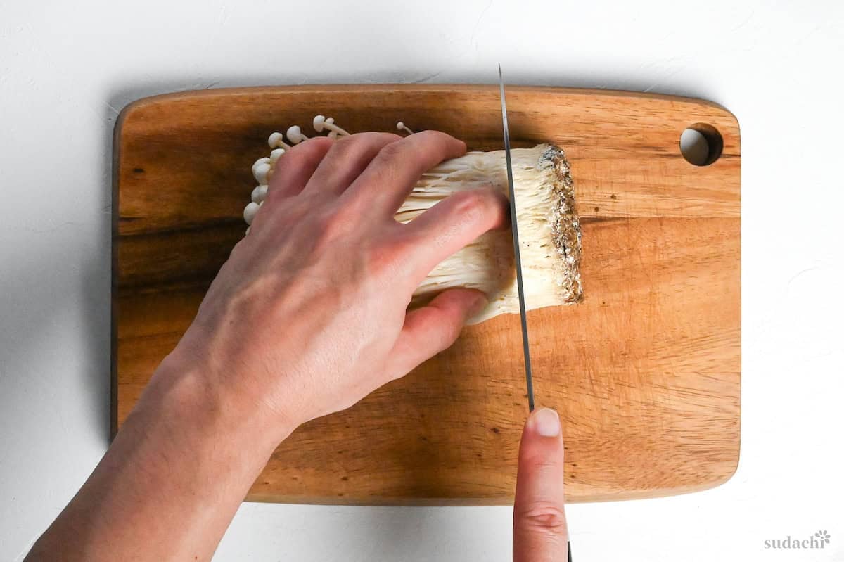 cutting the roots off of a bunch of white enoki mushrooms on a wooden cutting board