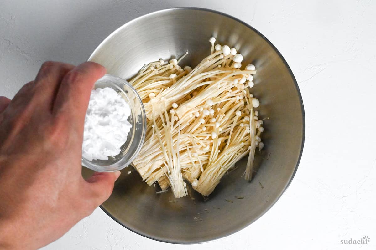 sprinkling potato starch over shredded enoki mushrooms in a mixing bowl