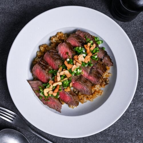 garlic fried rice topped with rare pan-fried beef steak slices, chopped green onions and crispy garlic chips in a white dish thumbnail