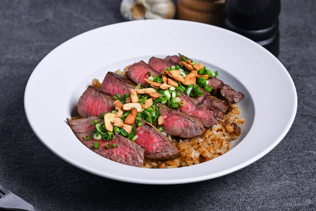 side view of garlic fried rice topped with rare pan-fried beef steak slices, chopped green onions and crispy garlic chips in a white dish