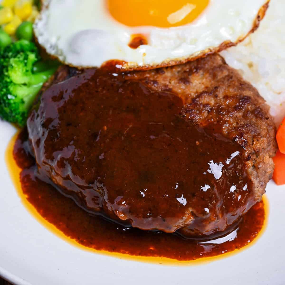 Japanese hamburg steak (hambagu) coated with homemade sauce on a white plate served with rice, vegetables and a fried egg thumbnail