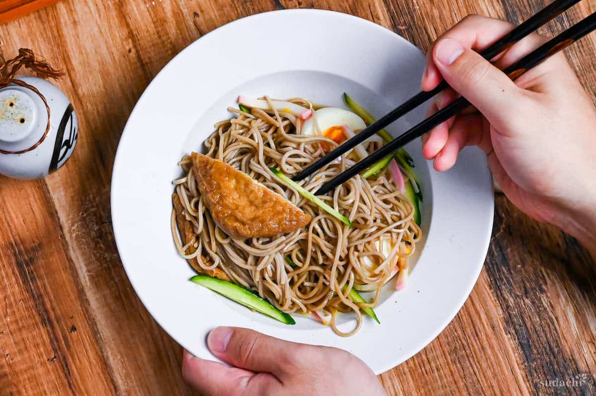 Hiyashi Kitsune Soba mixed in a white dish held with one hand, other hand holding black chopsticks