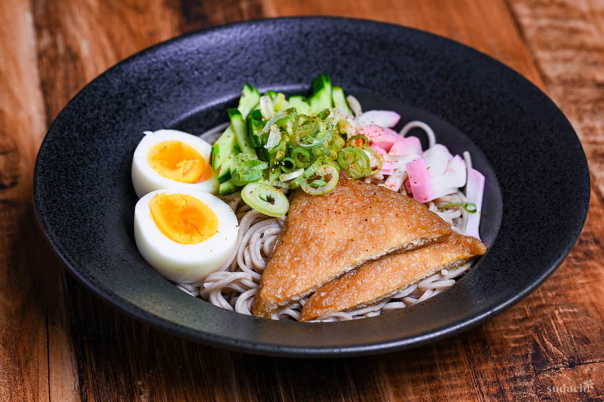 Hiyashi Kitsune Soba in a black dish topped with marinated tofu pouches, boiled eggs (halved), cucumber, kamaboko fishcakes, chopped green onions and shichimi togarashi side view