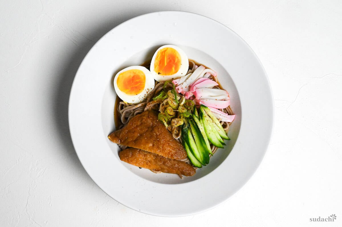 Hiyashi kitsune soba in a white dish topped with marinated tofu pouches, cucumber, chopped green onion, kamaboko fish cakes and boiled eggs