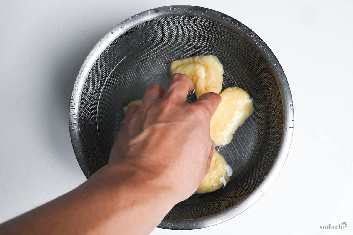 Four pieces of twice fried tofu pouches (aburaage) in a sieve over a mixing bowl filled with cold water