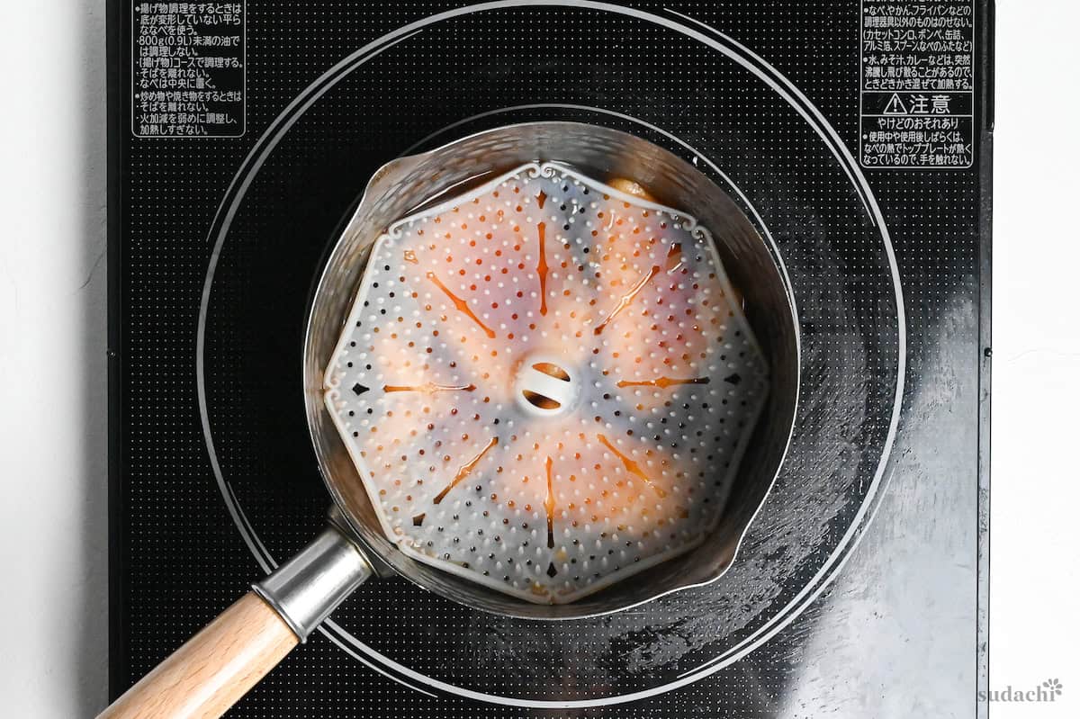Four pieces of twice fried tofu pouches (aburaage) in marinade in a pot on the stove topped with drop lid to weigh them down