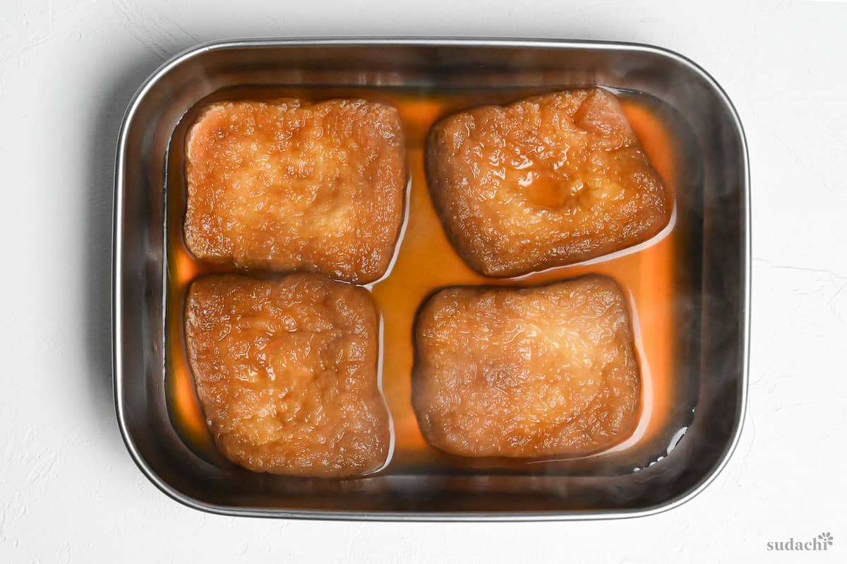 Four pieces of twice fried tofu pouches (aburaage) cooling in a container with marinade