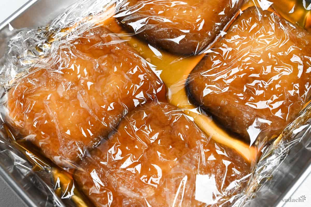 Four pieces of marinated twice fried tofu pouches (aburaage) in a container topped with plastic wrap close up
