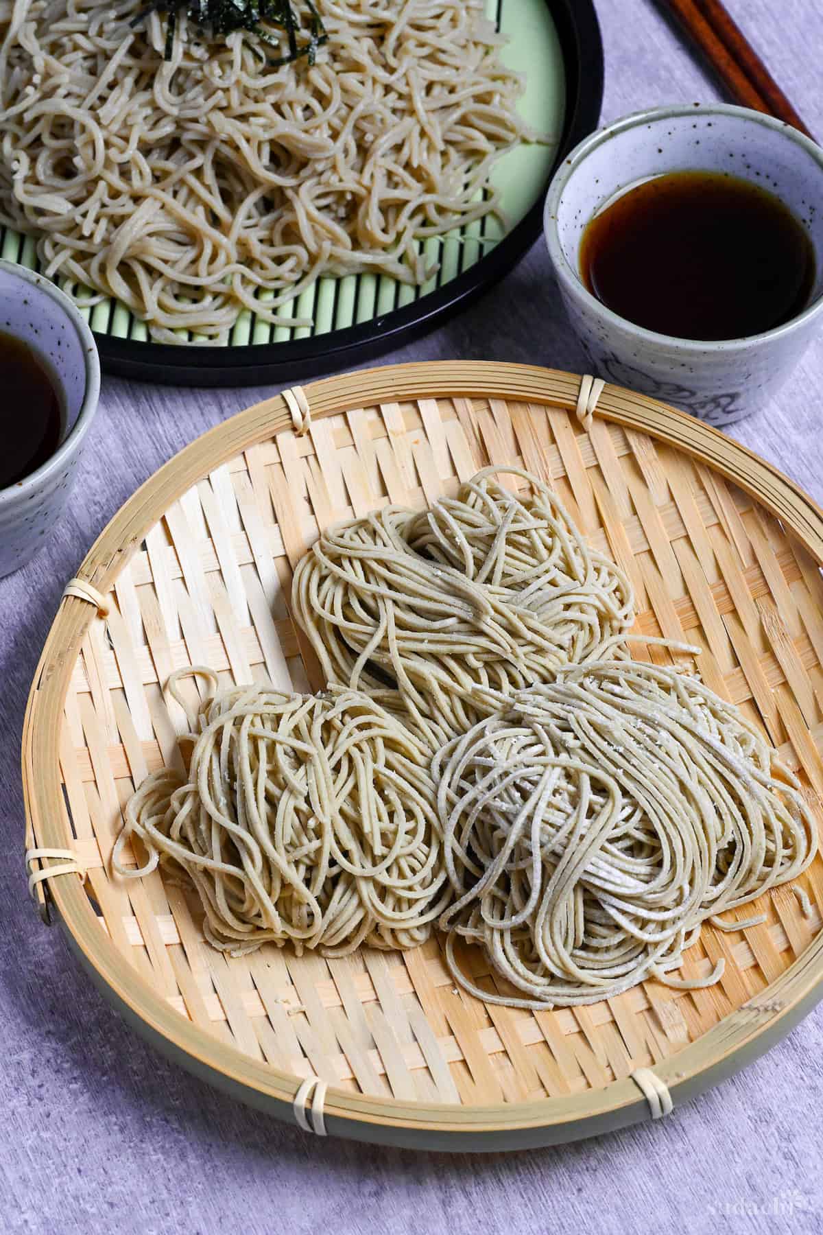 three bundles of homemade soba noodles on a bamboo tray with zaru soba and dipping sauce in the background