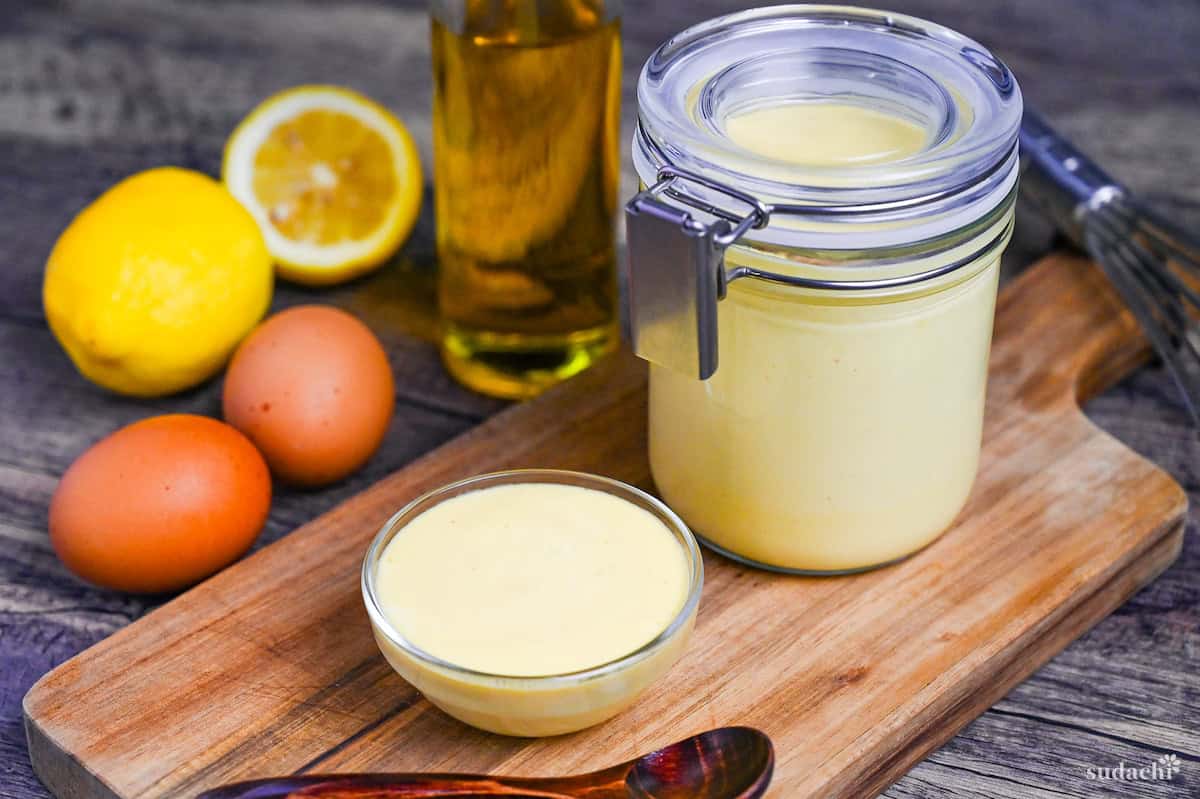 Homemade Japanese Mayonnaise (Kewpie Style) in a glass jar and glass bowl on a wooden chopping board with ingredients in the background
