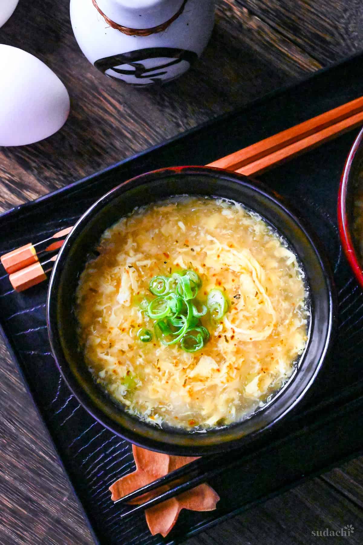 Kakitamajiru (Japanese egg drop soup) topped with chopped green onions and shichimi togarashi served in a black soup bowl on a black tray with wooden chopsticks