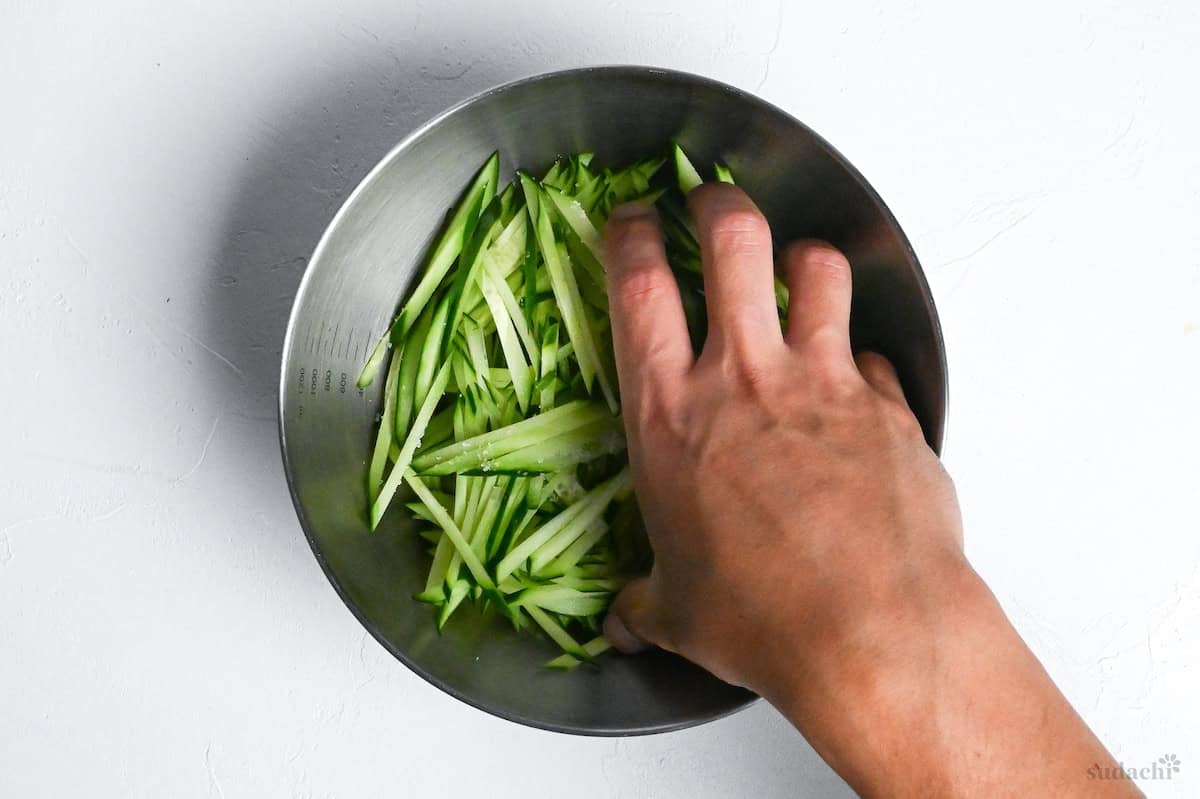 salting julienned cucumbers in a mixing bowl on a white background