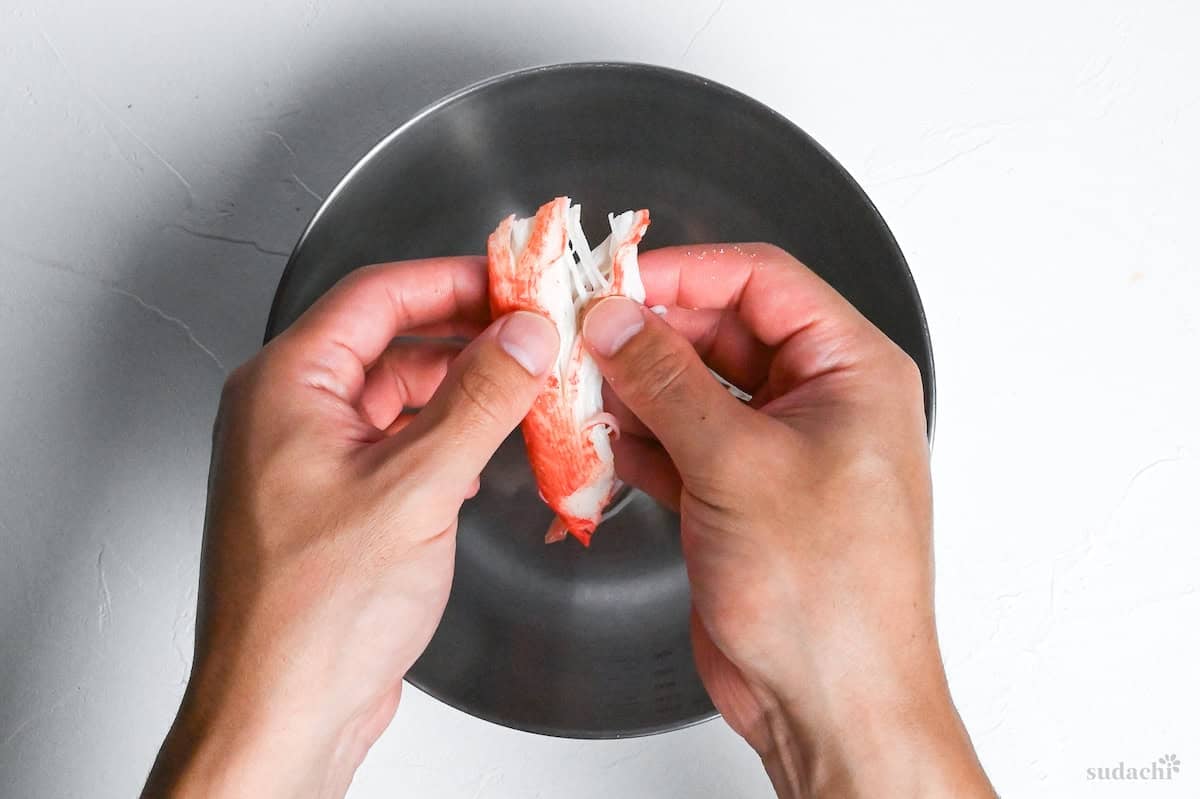 shredding imitation crab sticks by hand into a steel mixing bowl on a white background
