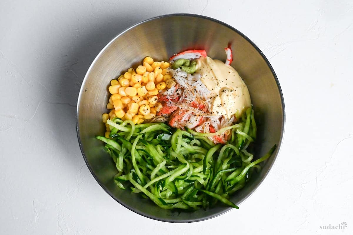 juleunned cucumber, canned corn, shredded imitation crab, mayo and seasonings in a steel mixing bowl on a white background