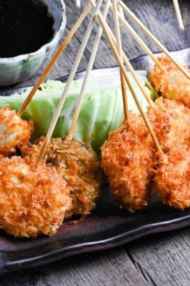 Kushi age/Kushi katsu battered skewers on a brown rectangular plate resting on a wedge of cabbage with dipping sauce in the background
