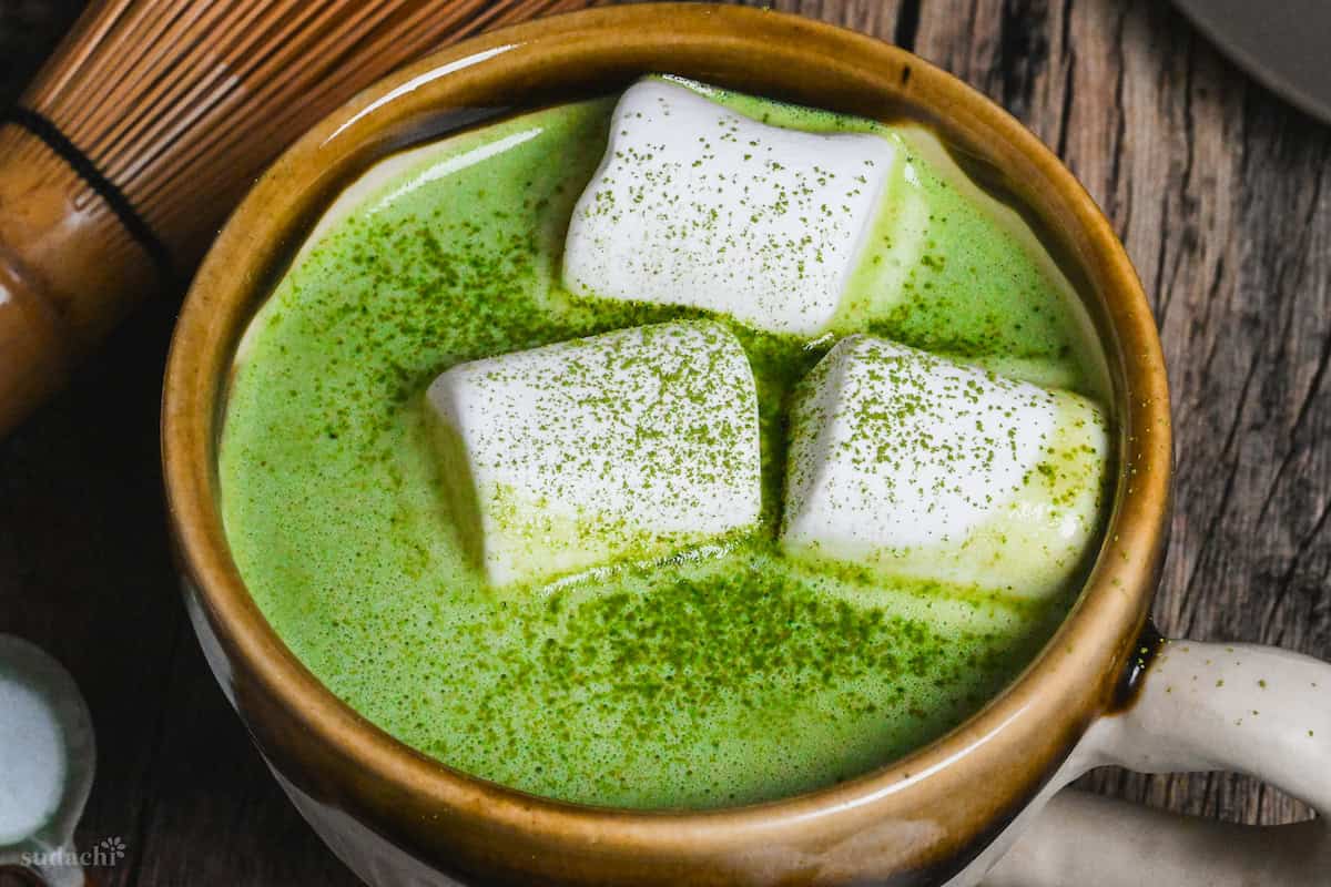 Matcha white hot chocolate in a brown and cream Japanese ceramic mug topped with marshmallows and matcha powder on a dark wooden background