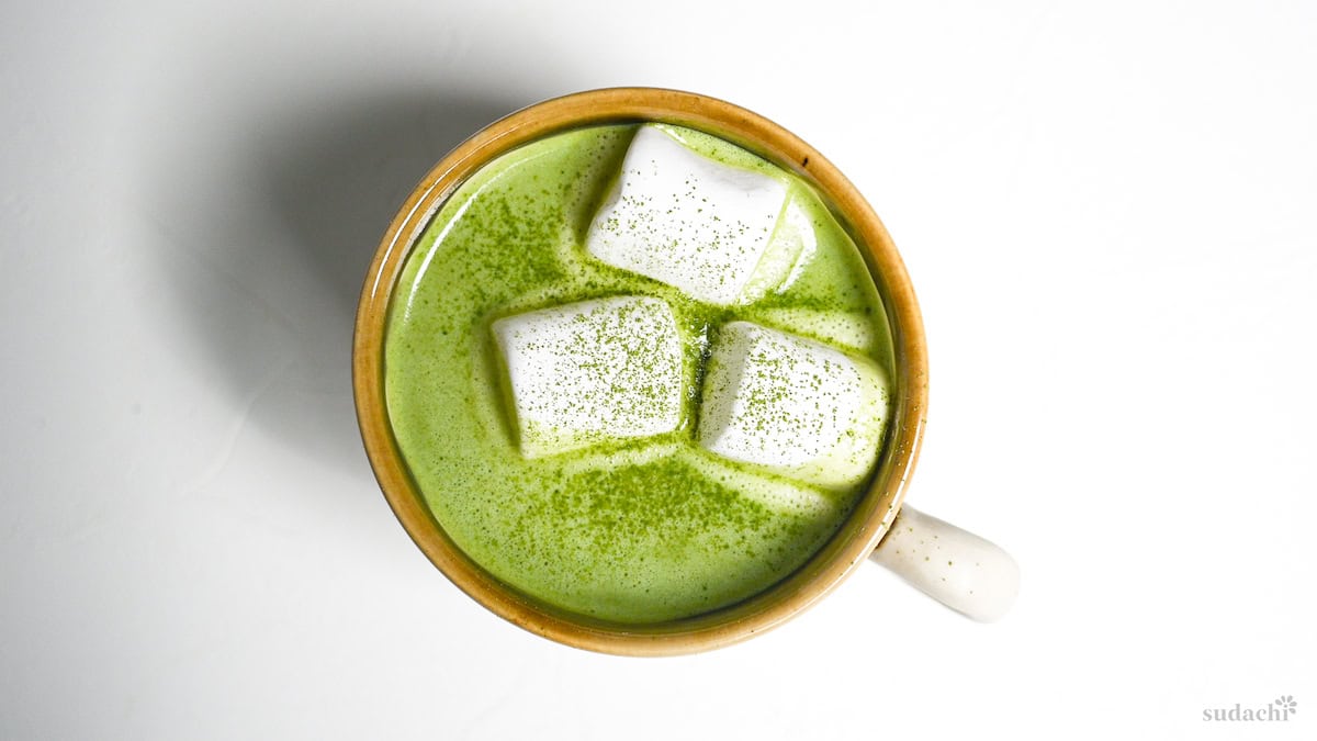 Matcha white hot chocolate in a brown and cream Japanese ceramic mug topped with marshmallows and matcha powder on a plain white background