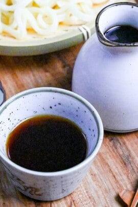 homemade mentsuyu (Japanese noodle dipping sauce) in a white dipping bowl next to a jug of sauce and a tray of udon and soba noodles
