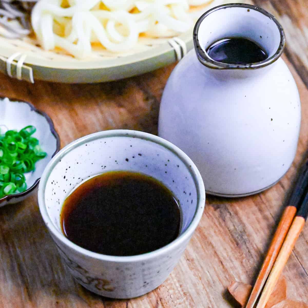 homemade mentsuyu (Japanese noodle dipping sauce) in a white dipping bowl next to a jug of sauce and a tray of udon and soba noodles