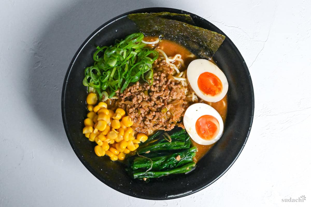 miso ramen topped with spicy ground pork, ramen eggs, blanched spinach, sweetcorn, chopped green onion and nori in a black bowl on a white background