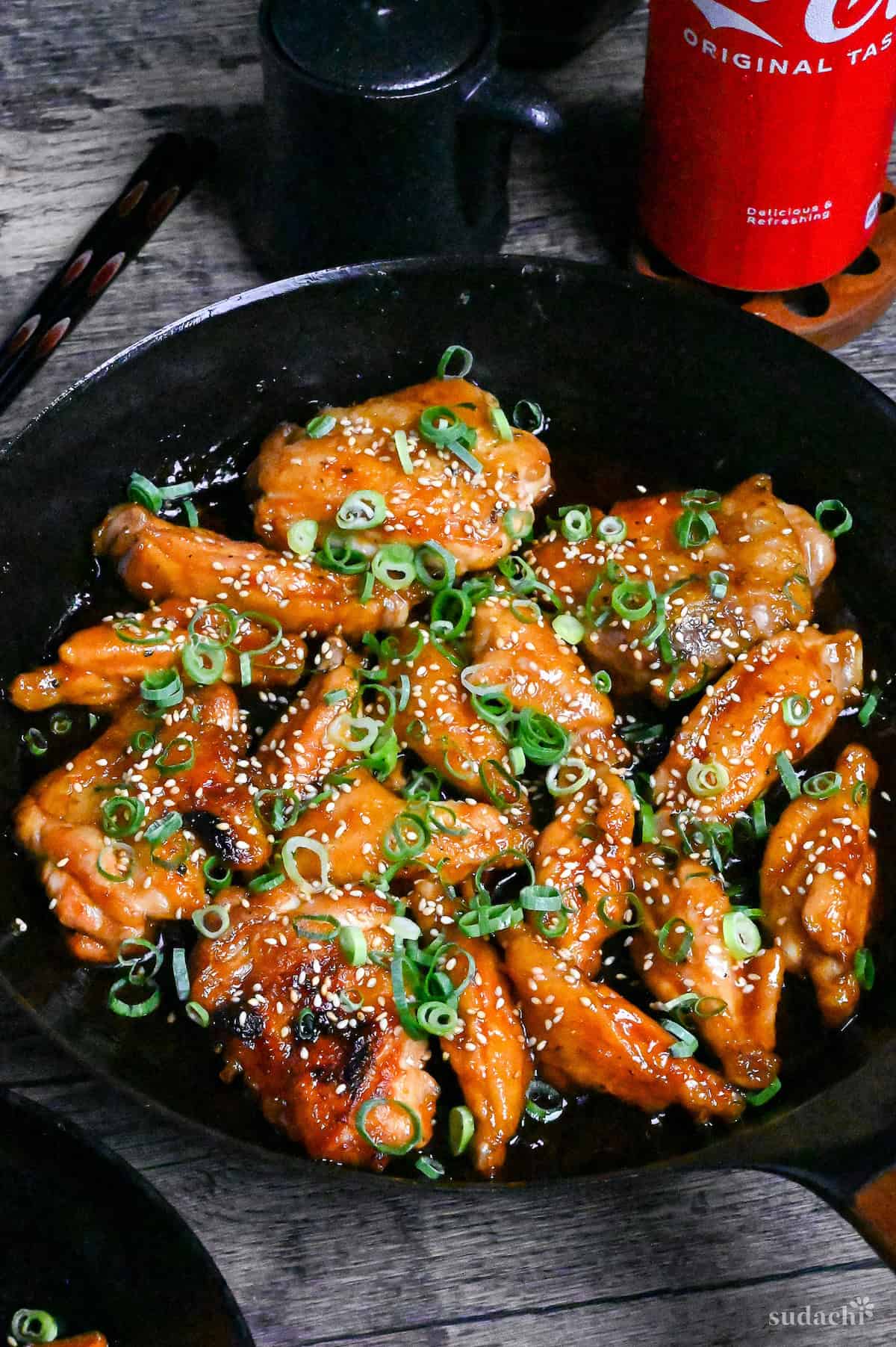 Mock teriyaki made with chicken wings in a black pan sprinkled with green onions and sesame seeds next to a ceramic soy sauce bottle and can of cola