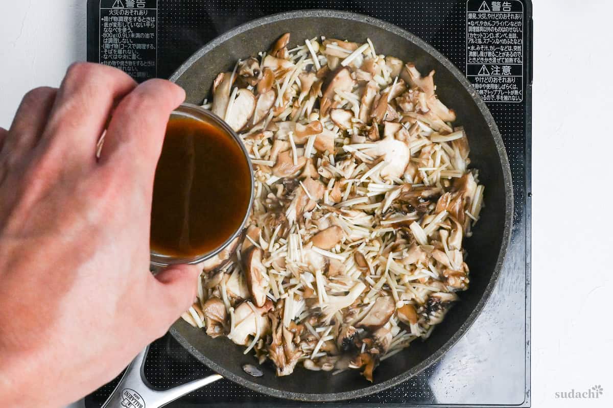 a hand pouring sauce over cooked mushrooms in a frying pan